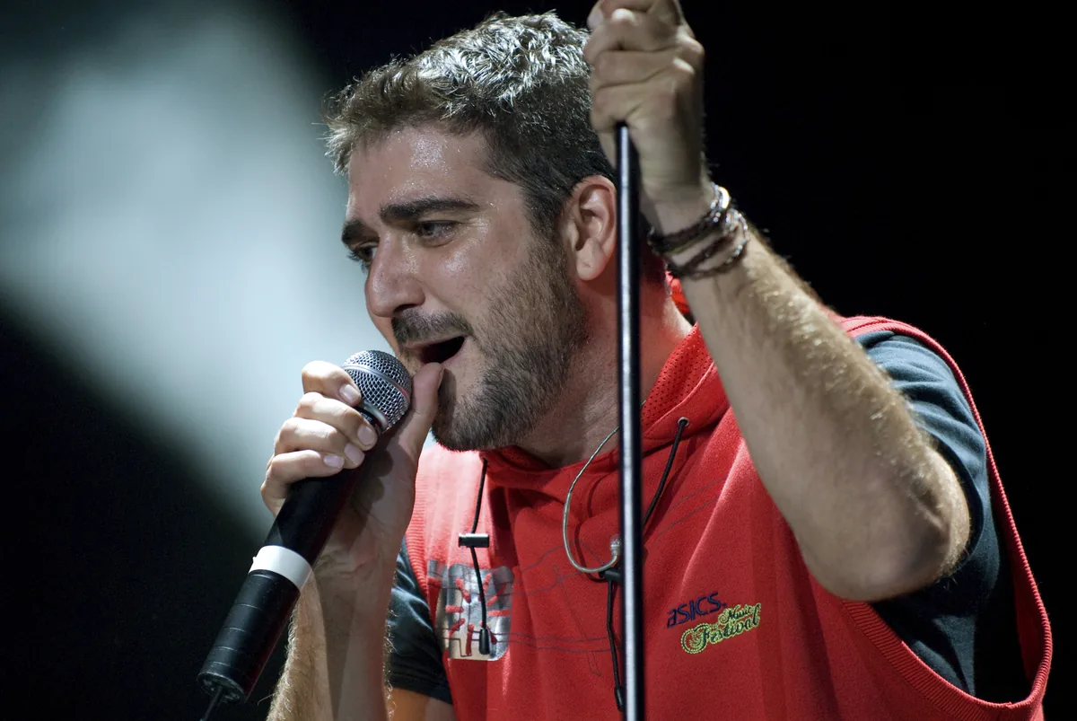 Antonio Orozco canta agarrado al pie del micrófono con una sudadera roja durante un concierto