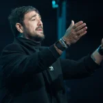 Antonio Orozco, de negro sobre un escenario oscuro, abre los brazos hacia el público mientras canta
