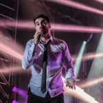 Melendi cantando en un concierto con focos de colores al fondo