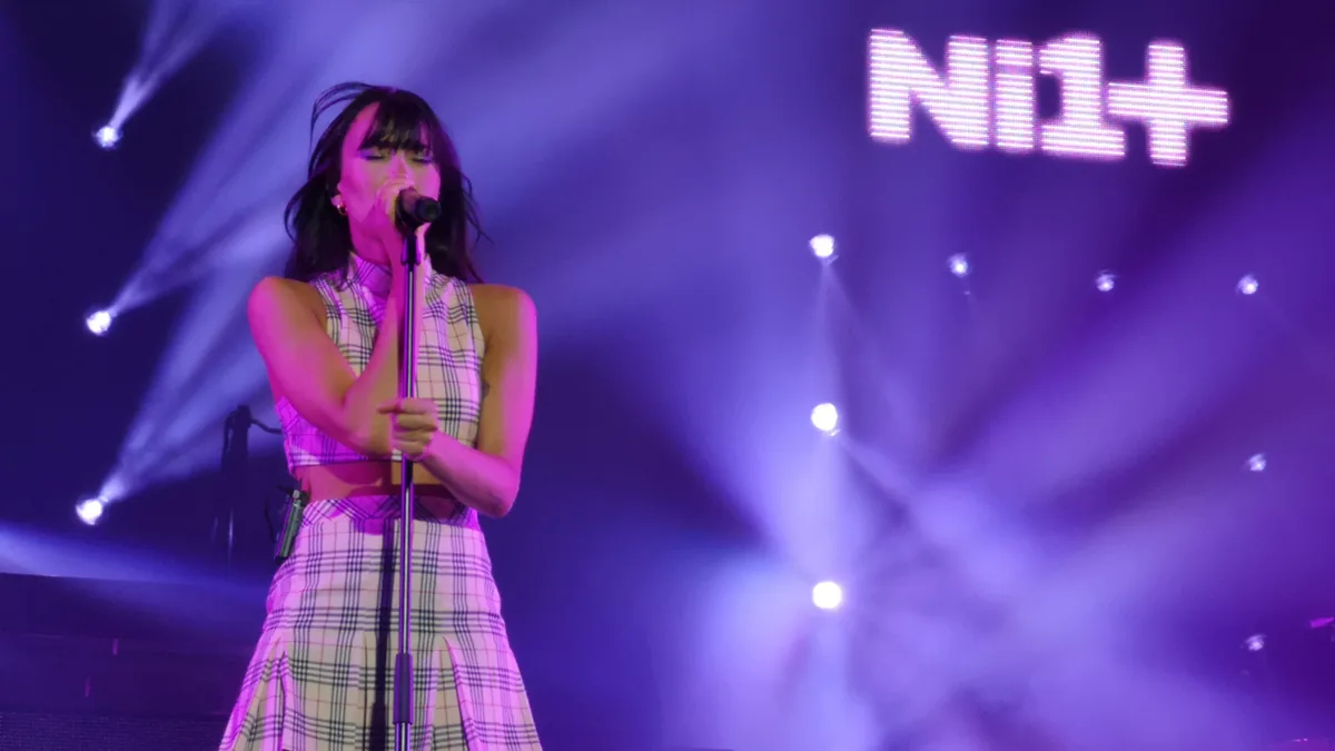 Aitana canta con el micrófono de pie bajo luces moradas en un concierto