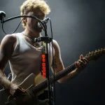 Dani Fernández canta y toca la guitarra eléctrica sobre el escenario, con camiseta blanca de tirantes
