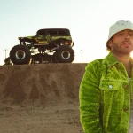Feid con chaqueta verde y gorro blanco junto a un monster truck