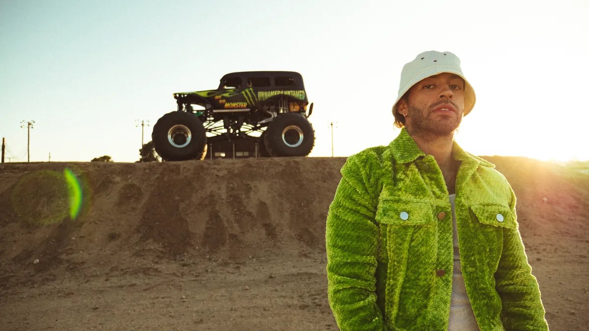 Feid con chaqueta verde y gorro blanco junto a un monster truck