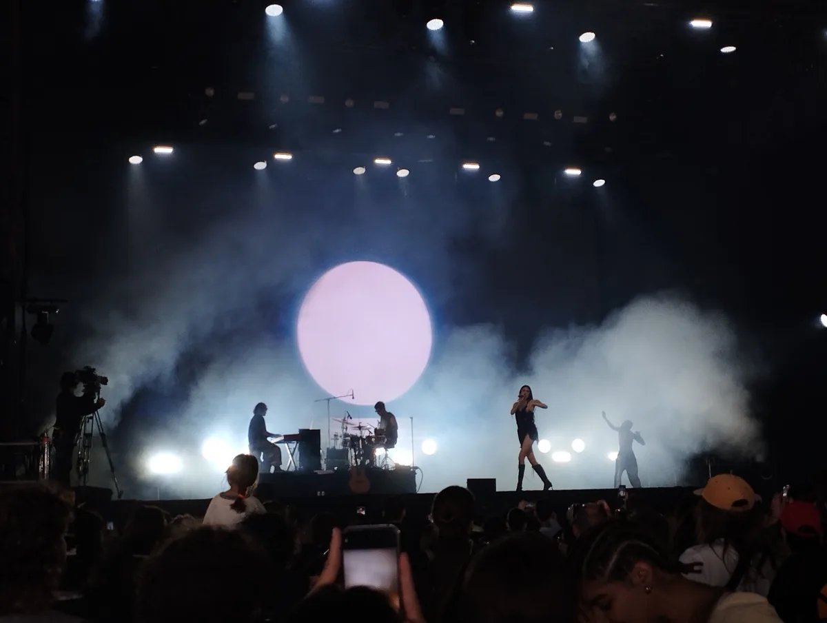 Judeline canta sobre el escenario ante un gran círculo de luz rosa, con su banda al fondo