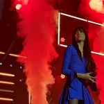 Aitana con vestido azul sobre el escenario, entre columnas de humo rojo