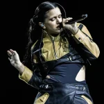 Rosalía canta con el micrófono en la mano y chaqueta de cuero negra y dorada