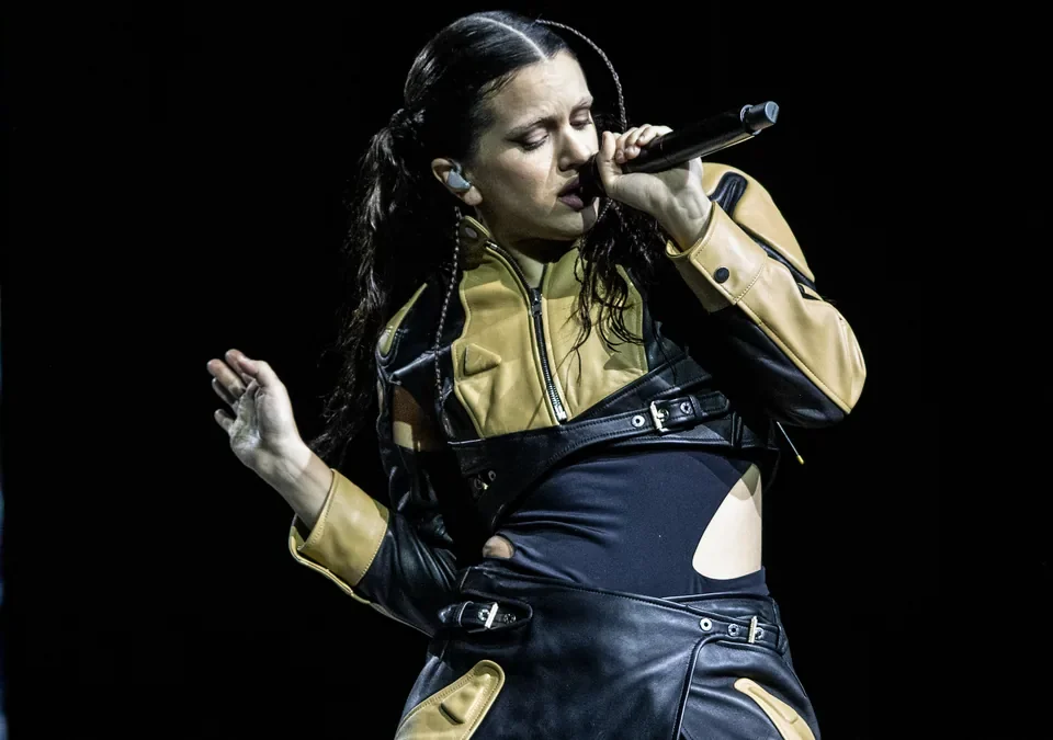 Rosalía canta con el micrófono en la mano y chaqueta de cuero negra y dorada