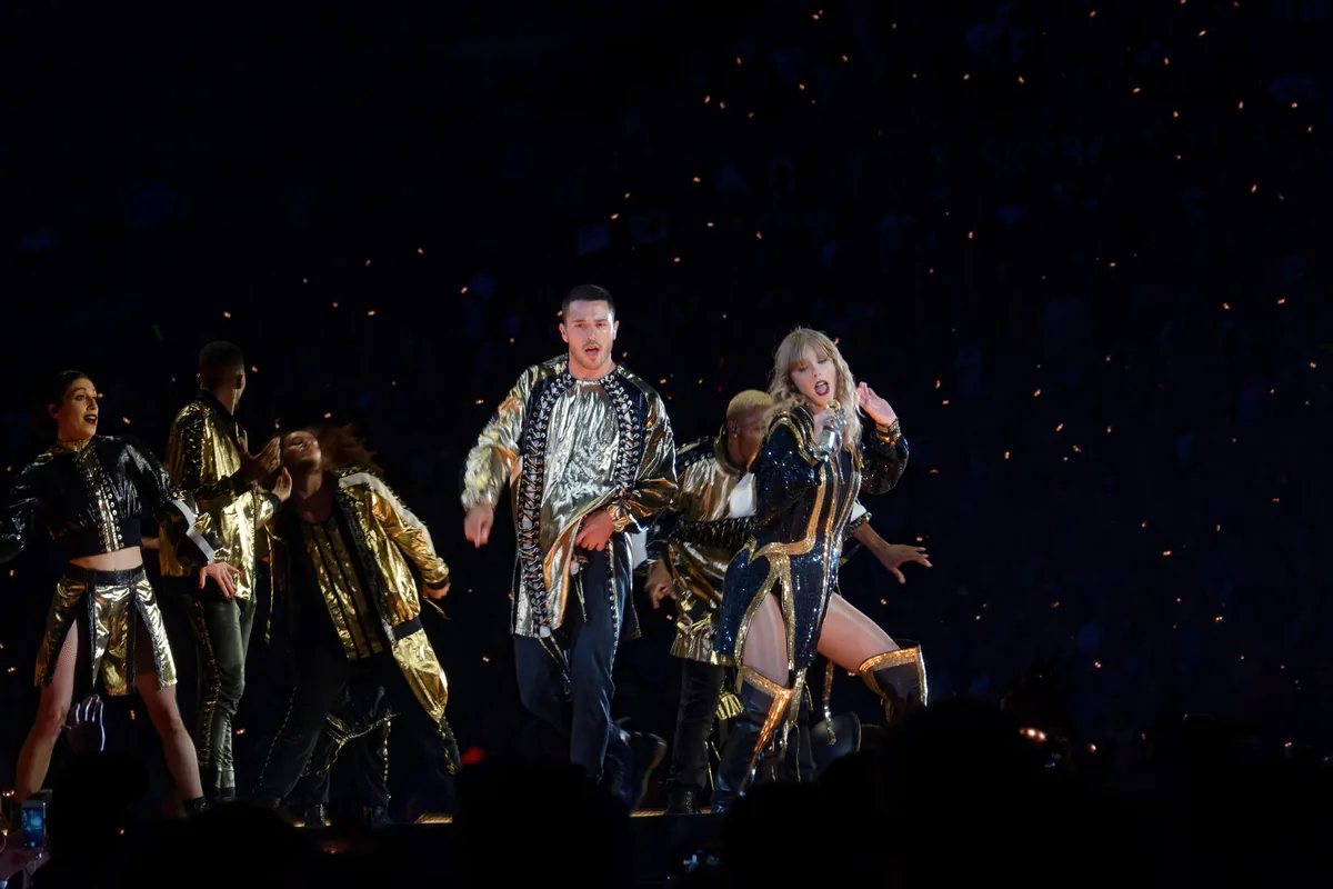 Taylor Swift con un traje de lentejuelas negro y dorado, rodeada de bailarines en el escenario